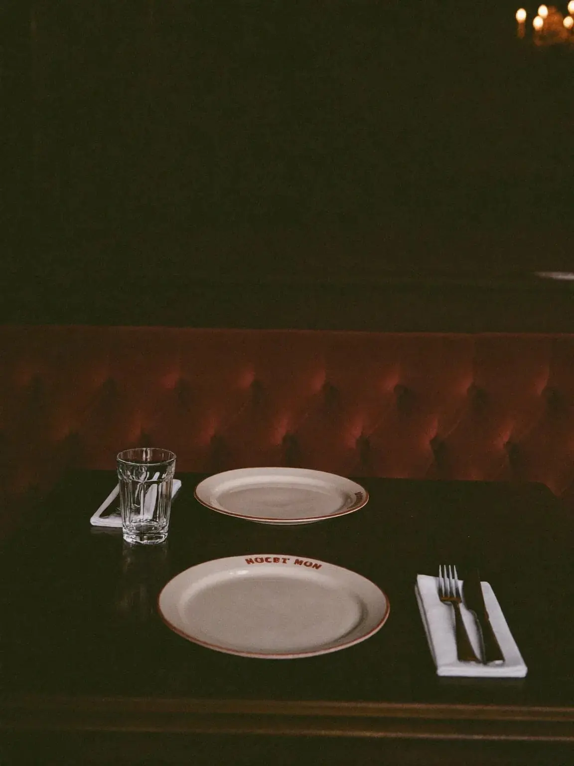 A laid table with plain white plates and folded napkin against a deep red banquette in the dining room.