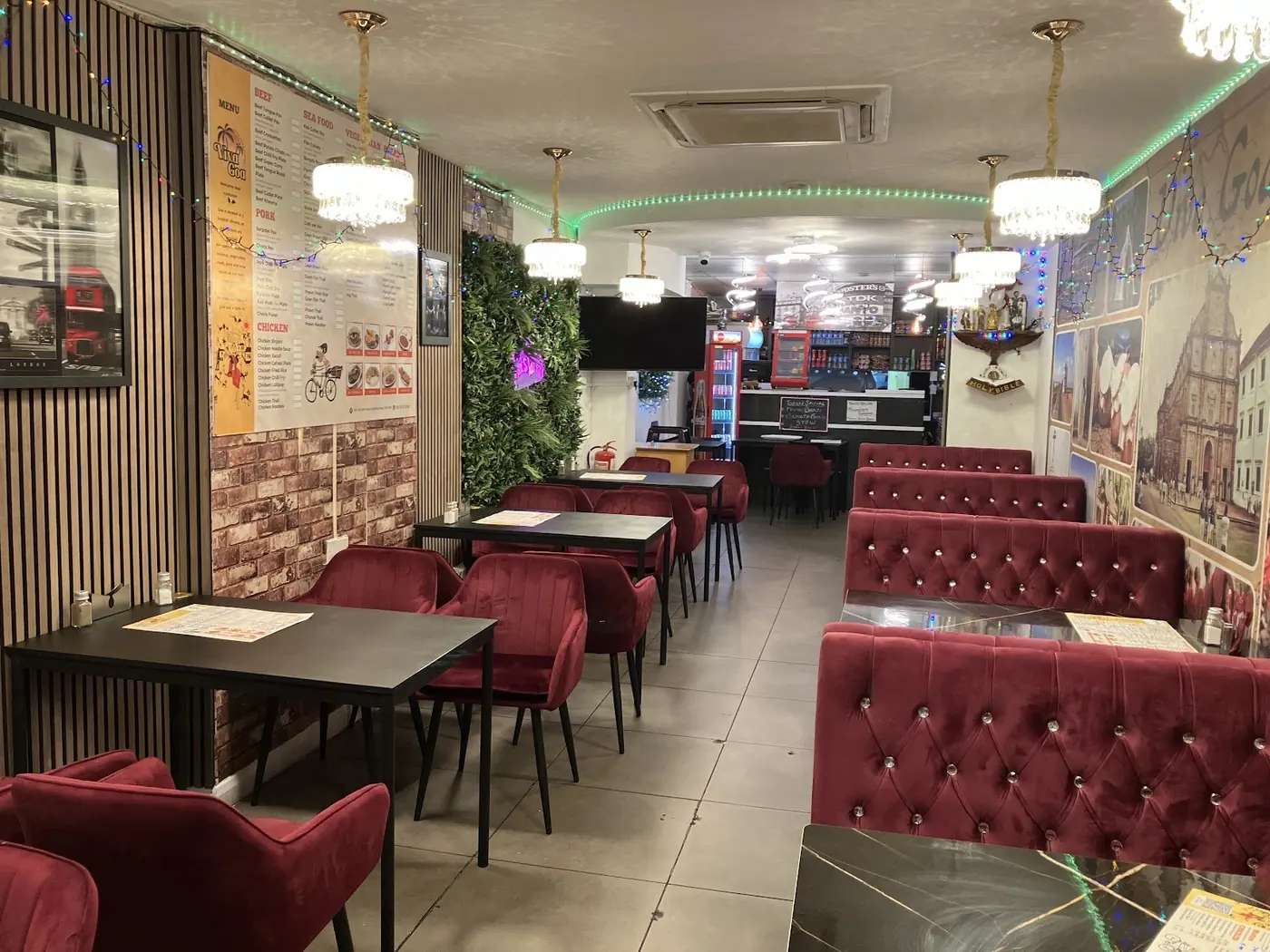 Interior of Velankanni Restaurant: a row of deep red tufted banquettes and dark tables along the wall, chandeliers and green neon ceiling trim, with the service counter at the back.