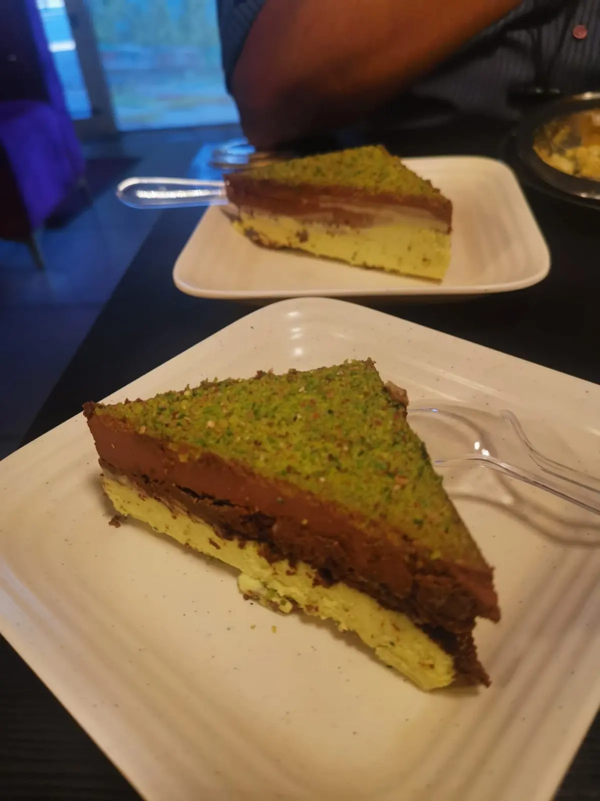 A wedge of layered chocolate and pistachio cake on a square white plate, on a dark table in dim blue-tinted light.