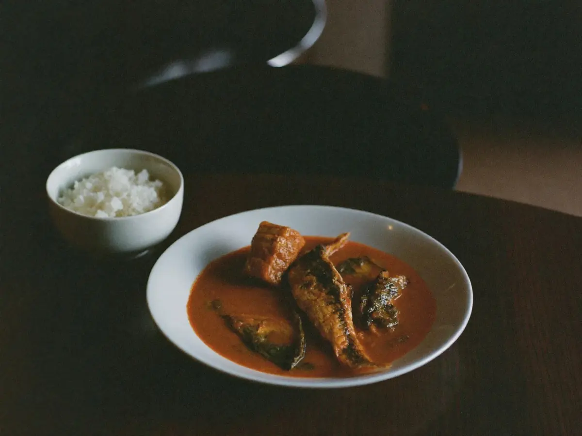 A Goan-style fish curry in a plain white bowl with rice alongside, on a dark table in low restaurant light.