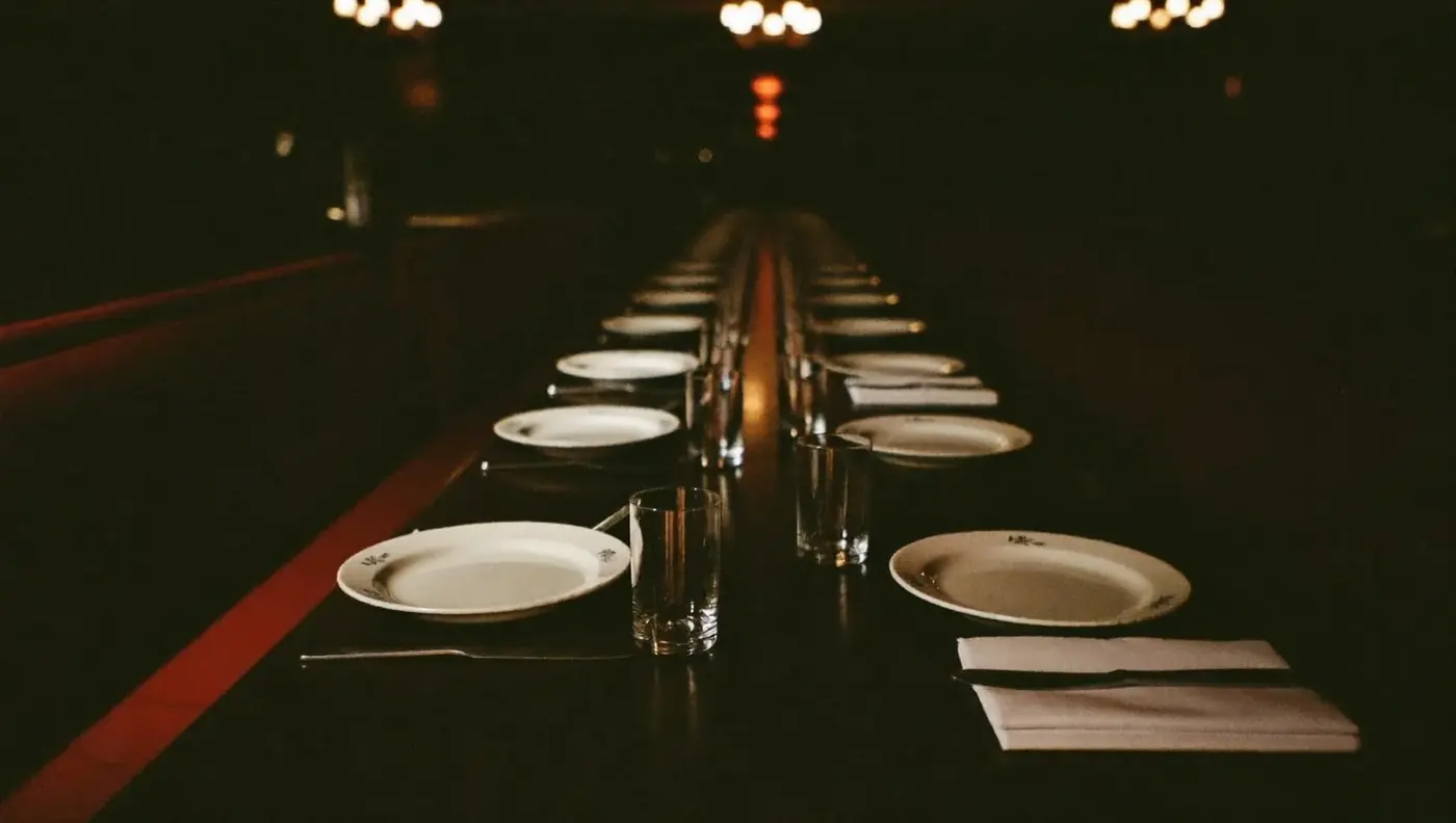 A long dining table set with plain white place settings running back into the warm low-lit dining room.